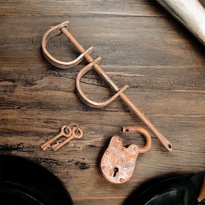 Puede incluir: Una vista cenital muestra objetos metálicos antiguos sobre una superficie de madera. La imagen incluye un candado oxidado, dos llaves y un par de grilletes. Los objetos tienen una apariencia desgastada y envejecida, lo que sugiere que son vintage o reutilizados.