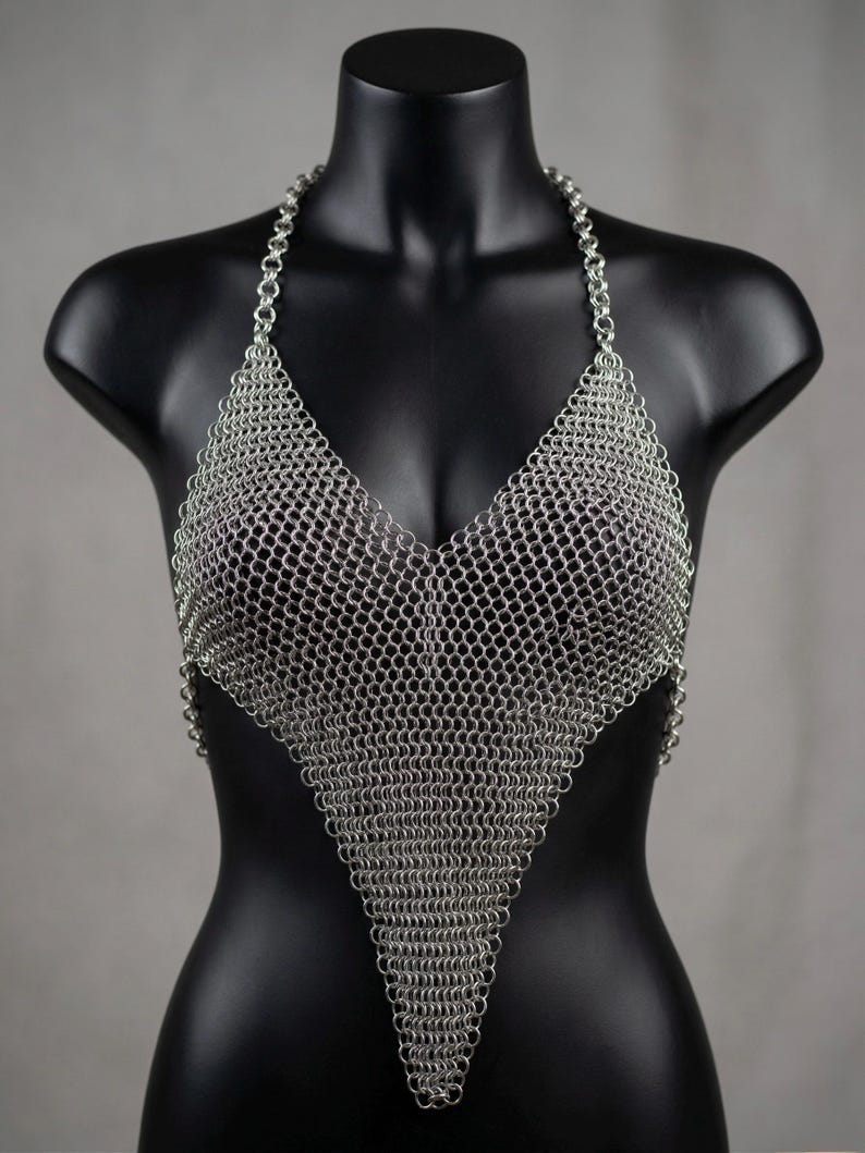 May include: A silver chainmail halter top displayed on a black mannequin. The top is constructed of small, interlocking metal rings, forming a triangular shape that covers the chest and a halter neck. The metal has a shiny, reflective quality.