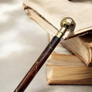 May include: A close-up of a decorative walking cane with a dark brown wooden shaft featuring gold-colored veining. The handle is gold-colored and topped with a textured, spherical design. The cane rests near open books.