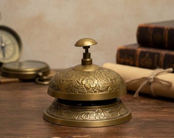 Op de afbeelding: Een vintage messing bel met bloemengravures. De bel heeft een koepelvormige bovenkant met een kleine handgreep en een uitlopende basis. De bel staat op een houten oppervlak, met een kompas, boeken en een rol in de achtergrond. De bel is ongeveer 10 cm hoog.
