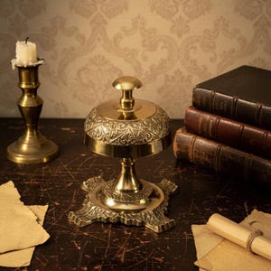 May include: A polished brass service bell with an ornate floral engraving on the dome and a square base with decorative feet. The bell features a central plunger button at the top and is constructed from a metallic material with a reflective finish.