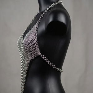 May include: A silver chainmail halter top displayed on a black mannequin. The top is made of small, interlocking metal rings, forming a V-neck design. The chainmail extends down the torso and has a chain that wraps around the back.