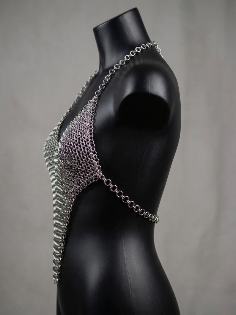 May include: A silver chainmail halter top displayed on a black mannequin. The top is made of small, interlocking metal rings, forming a V-neck design. The chainmail extends down the torso and has a chain that wraps around the back.