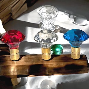 Puede incluir: Tres tapones decorativos para botellas con tapas facetadas en forma de joya en rojo, transparente y azul, cada uno sobre una base dorada. Descansan sobre una superficie de madera, con un vaso transparente y un objeto de vidrio verde en el fondo.