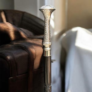 Könnte beinhalten: Silberfarbener Gehstock mit kunstvollem Griff und dunkelbraunem Schaft. Der Griff weist detaillierte Blumenmuster und eine geriffelte Oberseite auf. Der Stock steht vor einem braunen Ledersessel, wobei natürliches Licht die Szene erhellt.