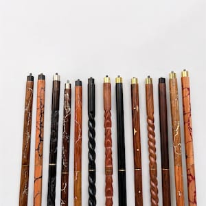 Puede incluir: Una colección de bastones de madera con varios diseños. Los bastones presentan diferentes vetas de madera, colores y elementos decorativos, incluyendo detalles dorados y patrones trenzados. Las puntas de los bastones tienen tapas metálicas.