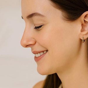 May include: A close-up profile of a person smiling, wearing a small gold hoop earring. The person has brown hair and is wearing a light-colored top. The background is a soft, neutral color.