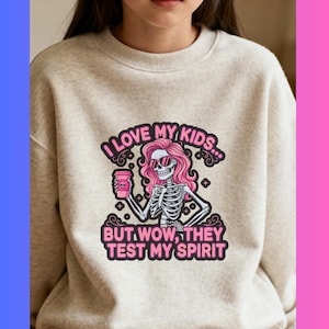 Può includere: Felpa beige con una grafica raffigurante uno scheletro con capelli rosa e occhiali da sole che tiene in mano una tazza di caffè. Il testo recita: "I LOVE MY KIDS... BUT WOW, THEY TEST MY SPIRIT."