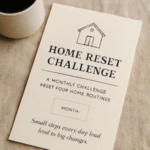 May include: A cream-colored card with the text "HOME RESET CHALLENGE" and a house illustration. The card includes prompts for a monthly challenge to reset home routines. A speckled ceramic mug with coffee sits nearby.