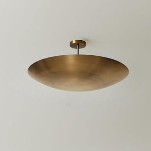 May include: A large, brass-colored ceiling light fixture. The fixture has a shallow, bowl-shaped shade and a small, circular mounting plate. The light hangs from a short rod. The overall design is minimalist and modern.