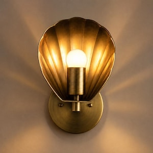 Op de afbeelding: Een wandlamp met een schelpvormige kap van messingkleurig metaal. Het armatuur bevat een centrale cilindrische fitting met een brandende gloeilamp, die een warm, stralend lichtpatroon op het muuroppervlak werpt.