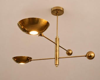 Könnte beinhalten: Eine moderne Messing-Pendelleuchte mit zwei schalenförmigen Leuchten und zwei kugelförmigen Akzenten. Die Leuchte wird an einer zentralen Stange aufgehängt und verfügt über verstellbare Arme, wodurch ein einzigartiges und stilvolles Lichtdesign entsteht.