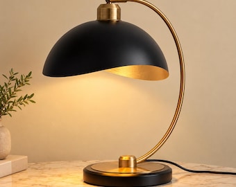 Könnte beinhalten: Eine Tischleuchte mit einem mattschwarzen, kuppelförmigen Schirm und goldfarbener Innenseite. Die Leuchte hat einen geschwungenen, goldfarbenen Metallarm, der mit einem runden Sockel verbunden ist, der außen schwarz ist und eine goldene Deckplatte hat.
