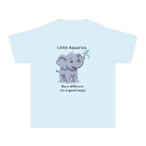 Op de afbeelding: Een lichtblauw t-shirt met een cartoonillustratie van een grijze olifant die water uit zijn slurf spuit. Boven de olifant staat de tekst Little Aquarius en daaronder de tekst Born different (in a good way) in zwarte letters.