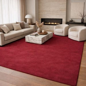 Peut inclure: Un grand tapis rouge uni recouvre le sol d'un salon. La pièce comprend un canapé beige, deux fauteuils crème et une table basse en marbre. Une cheminée avec un cadre noir et un feu allumé est en arrière-plan.