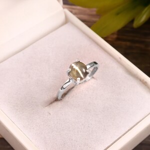 May include: A silver ring with a light brown, oval-shaped gemstone. The gemstone is set in a four-prong setting. The ring is displayed in a white jewelry box with a pink interior. The ring is a piece of jewelry.