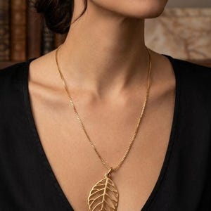 May include: A gold-colored necklace featuring a leaf pendant. The pendant has a detailed, openwork design, and hangs from a delicate chain. The necklace is worn against a black top.