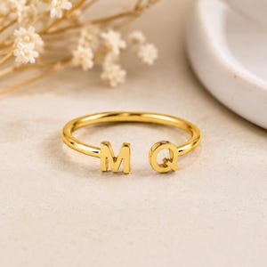Puede incluir: Un anillo abierto de tono dorado que presenta dos letras mayúsculas, M y Q, situadas en los extremos de la banda. El anillo tiene un acabado metálico liso y pulido, con un diseño circular sencillo que permanece abierto entre las letras.