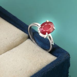 Puede incluir: Un anillo de plata con una gran gema de rubí de talla ovalada. El rubí es de color rojo intenso y está engastado en una montura de cuatro garras. El anillo se muestra en un joyero de terciopelo azul sobre un fondo turquesa.