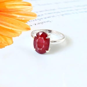 Puede incluir: Un anillo de plata con una gran gema de rubí de talla ovalada. La piedra roja vibrante es el punto focal, engastada en un engaste clásico de cuatro puntas. El anillo se muestra sobre una superficie blanca, con una flor naranja borrosa en el fondo.