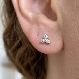 May include: A close-up of a small, silver stud earring featuring three clear, round gemstones in a triangular arrangement. The earring is set against the skin of an ear, with the person's hair visible in the background.