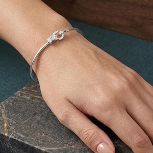 Può includere: Un braccialetto color argento con un design a nodo. Il braccialetto presenta una fascia sottile e un dettaglio centrale a nodo ornato da piccole pietre scintillanti. Il gioiello è esposto su una mano.