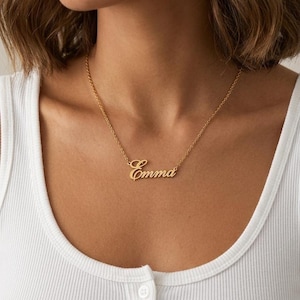 May include: A gold-colored necklace with the name "Emma" in a cursive font. The necklace is worn around the neck, resting on a white tank top. The chain is delicate, and the nameplate is centered.