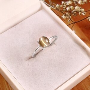 Puede incluir: Un anillo de plata con una piedra preciosa ovalada de color marrón claro. El anillo está engastado en un engaste de cuatro puntas y se presenta en una caja de joyería blanca. La banda del anillo es fina y pulida.