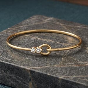 Può includere: Un delicato bracciale rigido in oro con due piccoli diamanti scintillanti e un esclusivo design a nodo. Il bracciale è esposto su una superficie di pietra grigio scuro, che ne esalta l'elegante semplicità. Un gioiello raffinato.