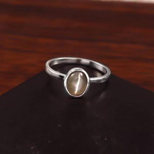 May include: A silver ring with an oval, light-colored gemstone. The ring band is thin and polished, and the gemstone is set in a simple bezel. The ring is displayed on a dark surface, showcasing its elegant design.