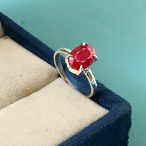 May include: A silver ring featuring a large, oval-cut ruby gemstone. The ruby is a vibrant red color and is set in a prong setting. The ring is displayed in a blue velvet jewelry box against a teal background.