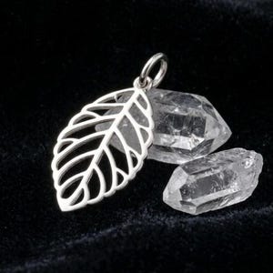 May include: A silver leaf pendant with intricate vein detailing, displayed on a black velvet surface next to two clear quartz crystals. The pendant has a small loop for a chain.