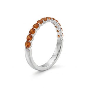 May include: A silver ring featuring a row of small, round, orange gemstones. The gemstones are set closely together, creating a continuous band of color. The ring has a polished, reflective surface.
