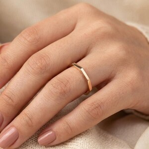 May include: A gold-colored ring with a geometric design. The ring has a series of straight edges and angles, creating a modern and minimalist aesthetic. The ring is worn on a hand with polished nails.