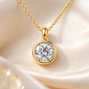 Könnte beinhalten: Eine goldene Halskette mit Anhänger und einem großen, runden, klaren Edelstein. Der Edelstein ist in einer goldenen Lünette gefasst und hängt an einer zarten Goldkette. Die Halskette wird auf cremefarbenem Satin präsentiert.