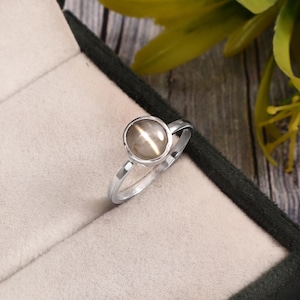 Puede incluir: Un anillo de plata con una gema redonda de color marrón claro. El anillo tiene un diseño sencillo y elegante, y se muestra en una caja de joyería. La gema tiene un efecto chatoyant, reflejando la luz de una manera única.