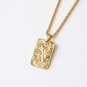 Puede incluir: Collar con colgante rectangular dorado con superficie texturizada y cadena a juego. El colgante tiene un diseño rugoso y orgánico. El collar se muestra sobre un fondo blanco.
