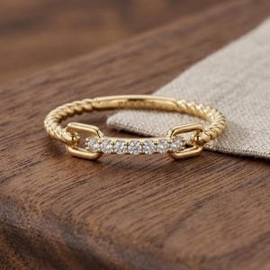 May include: A gold ring with a twisted band and a row of small, clear stones set in a rectangular design. The ring is on a wooden surface with a neutral-colored fabric in the background.