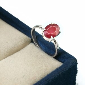 May include: A silver ring with a large, oval-cut ruby gemstone. The ruby is a deep red color and is set in a four-prong setting. The ring is displayed in a blue velvet jewelry box.