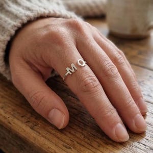May include: A silver ring featuring the letters M and Q as the focal point of the band. The letters are connected in a continuous line, forming the top section of the ring. The metal has a polished finish, reflecting light across the surface.