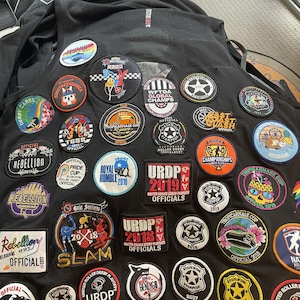 Könnte beinhalten: Schwarze Weste mit zahlreichen bunten Aufnähern. Aufnäher zeigen Texte wie "Rebellion", "URDP" und "Tropicarnage Cup". Designs umfassen Sterne, Derby-Skater und Event-Logos. Die Weste ist wahrscheinlich für einen Roller-Derby-Offiziellen.