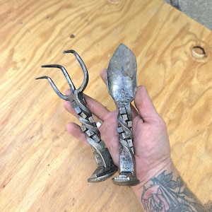 May include: Hand-forged metal garden tools, a small rake and a trowel, with twisted handles and decorative bases. The tools are made of dark metal and are displayed on a light wood surface.