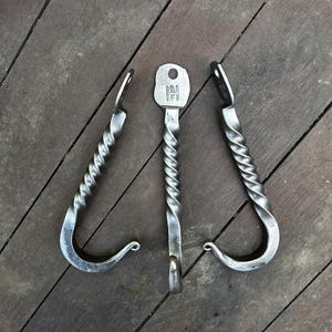 May include: Three hand-forged metal hooks with a twisted design. The hooks are silver in color and have a key-shaped center hook. The hooks are displayed on a wooden surface.
