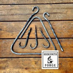 May include: Hand-forged metal hooks and a triangular frame rest on a wooden surface. The hooks have a curved design, and some feature a twisted pattern. A square logo with the text "HOOKSMITH FORGE" is also present.
