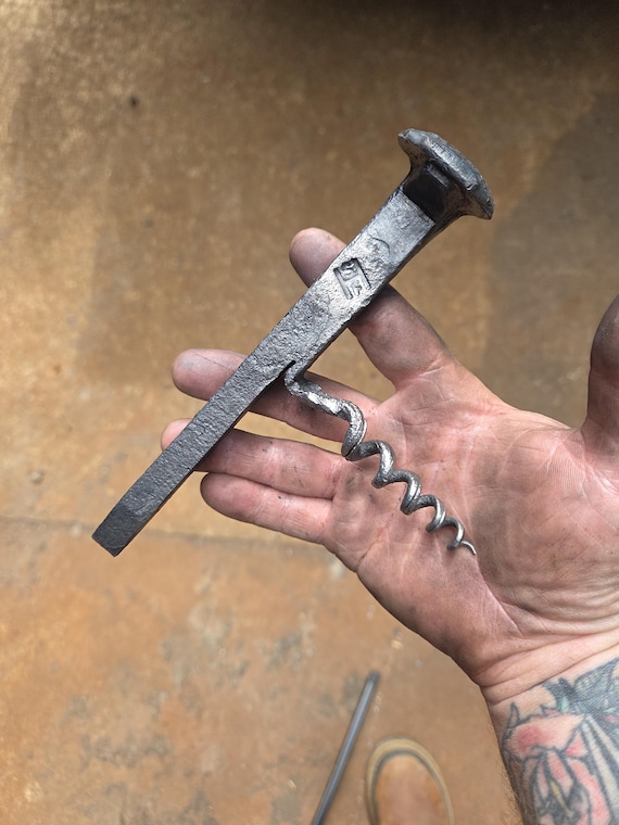 A real railroad spike transformed by a blacksmith into a dual-function corkscrew and bottle opener — heavy, sculptural, and genuinely one-of-a-kind bar art.