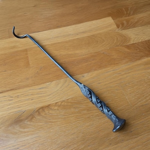 May include: A hand-forged, dark gray metal fire poker with a curved hook at one end and a decorative twisted handle. The poker is designed for use in a fireplace or fire pit. The handle has a textured, woven design.