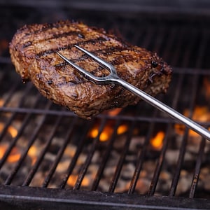May include: A grilled steak with grill marks is held by a silver fork over a charcoal grill with flames. The steak is a rich brown color, and the grill is black with glowing orange flames.