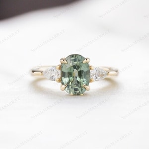 Könnte beinhalten: Ein goldener Ring mit einem großen, oval geschliffenen, hellgrünen Edelstein im Zentrum. Zwei kleinere, birnenförmige, klare Edelsteine flankieren den Mittelstein. Der Ring hat ein zartes Band und ein klassisches Drei-Steine-Design.