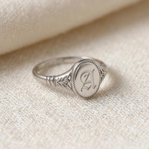 Könnte beinhalten: Ein silberner Siegelring mit einem ovalen Gesicht, das mit dem Buchstaben "A" graviert ist. Der Ring hat ein verziertes, detailliertes Band mit einem dekorativen Muster. Der Ring wird auf einer strukturierten, cremefarbenen Oberfläche gezeigt.