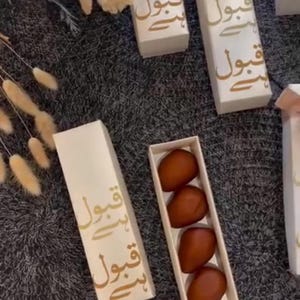 Pu&ograve; includere: Diverse scatole regalo rettangolari bianche con scritta araba dorata sono disposte su una superficie grigia. Una scatola aperta rivela tre dolcetti marroni di forma ovale.